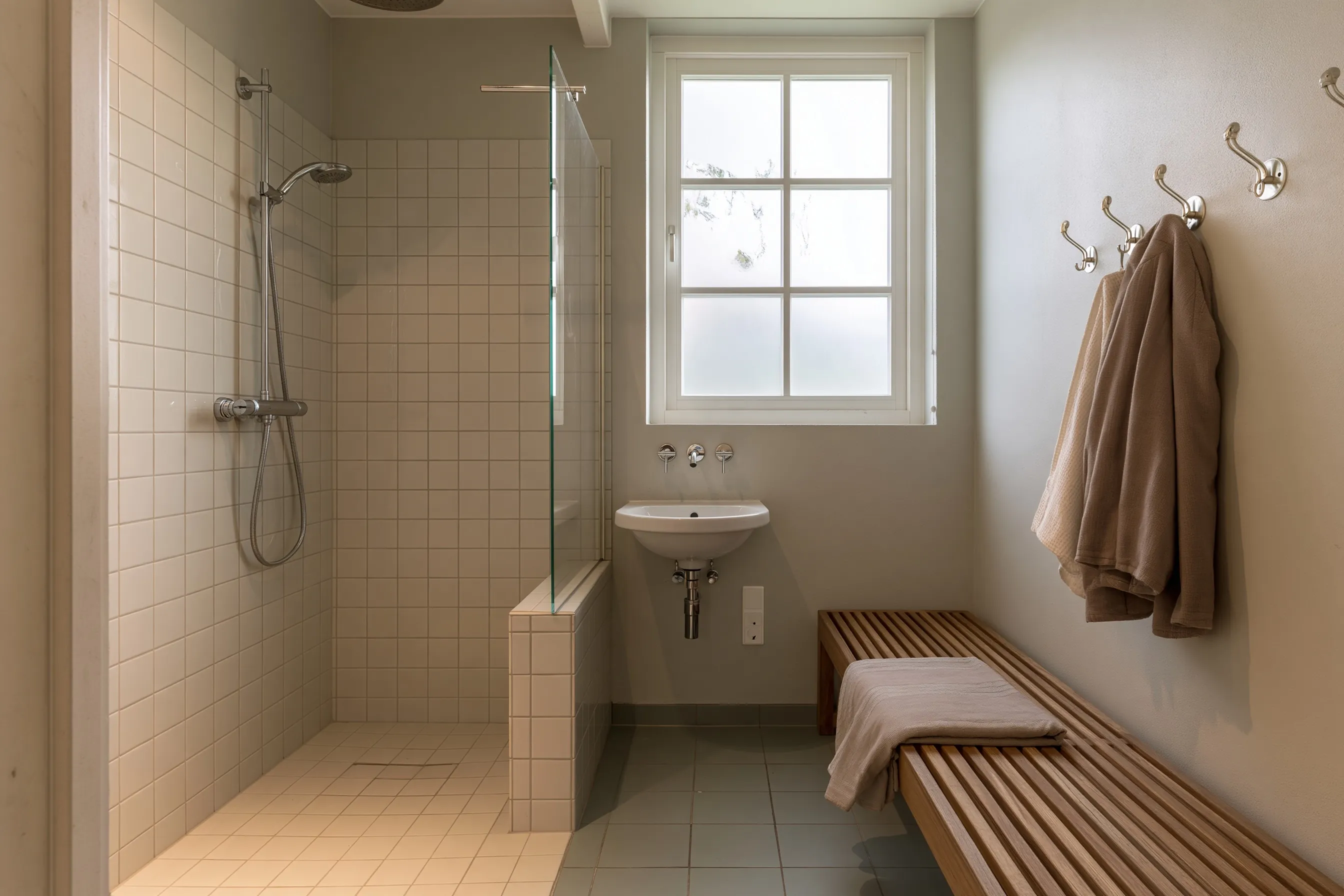 Pool house shower room with a tiled walk-in shower behind a glass panel, a wall basin under a frosted window, a slatted bench and robe hooks